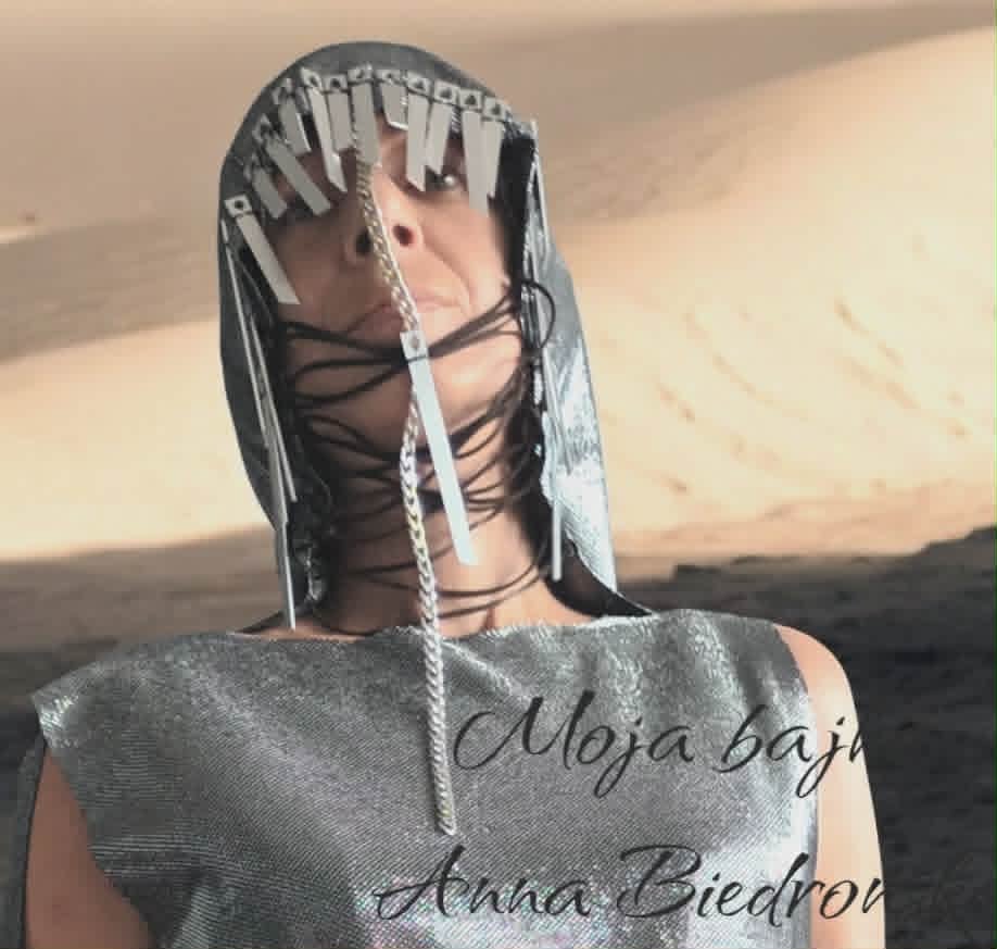 Person wearing metallic gray sleeveless top with elaborate beaded head covering and chains, standing in desert landscape
