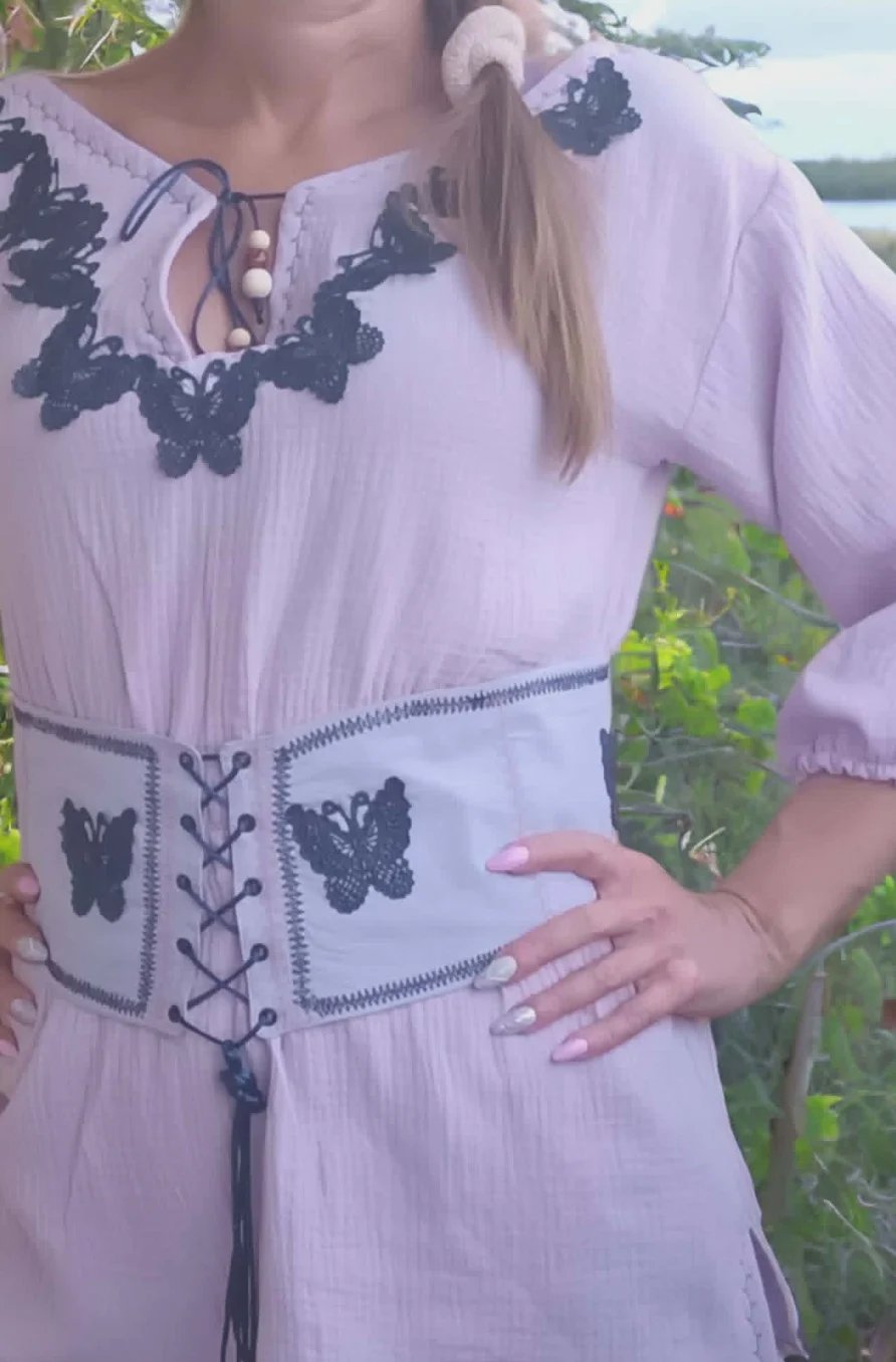 Person wearing lavender dress with black butterfly embroidery and corset-style waist detail, outdoors with greenery in background