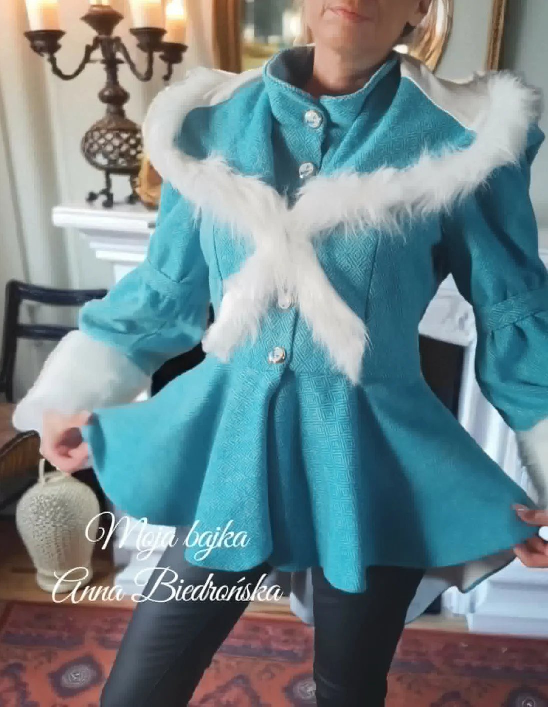 Woman wearing a turquoise coat with white faux fur collar and cuffs, posing indoors with candlabra in background
