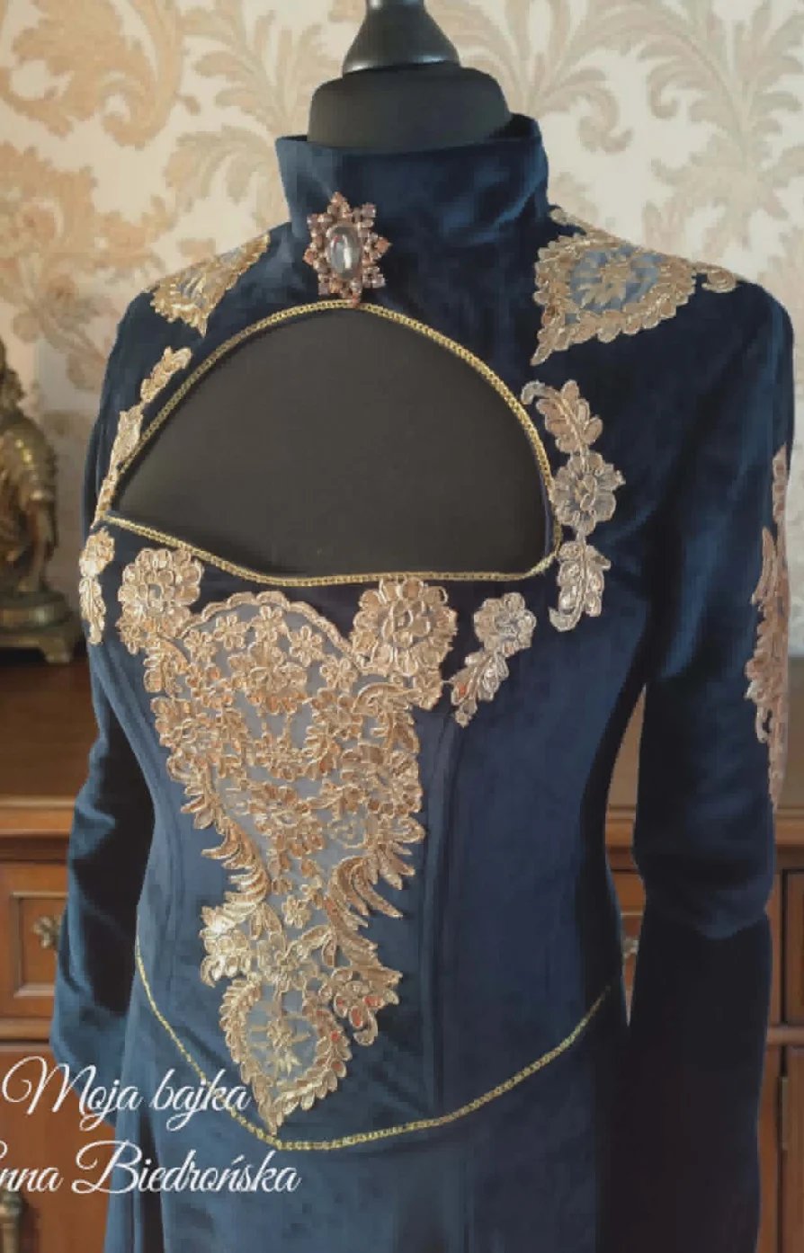 Mannequin displaying dark blue velvet jacket with ornate gold embroidery and beading on a patterned wallpaper background