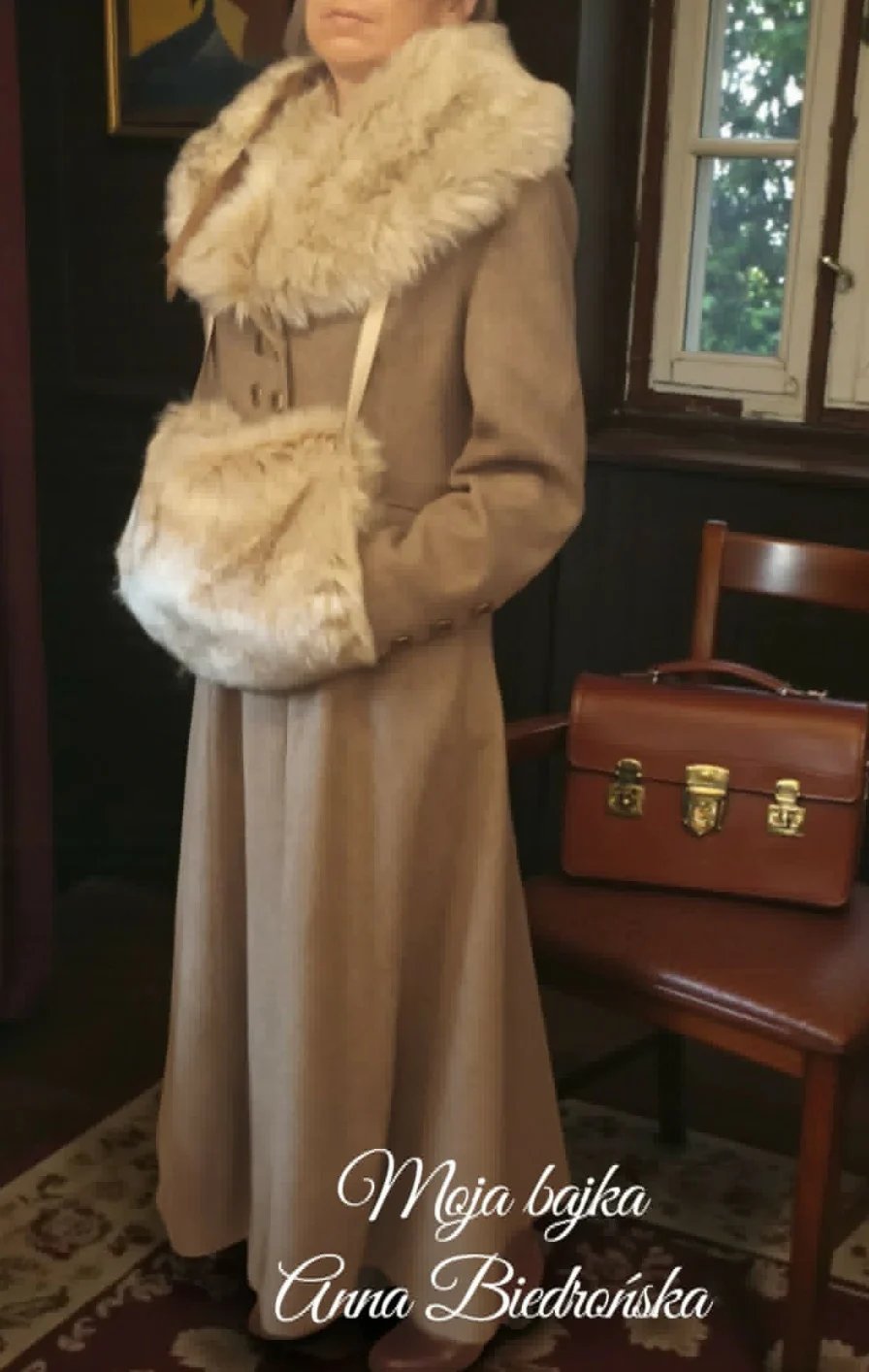 Woman wearing a beige coat with fur collar and cuff, holding a matching fur muff, standing indoors next to a wooden chair and leather briefcase