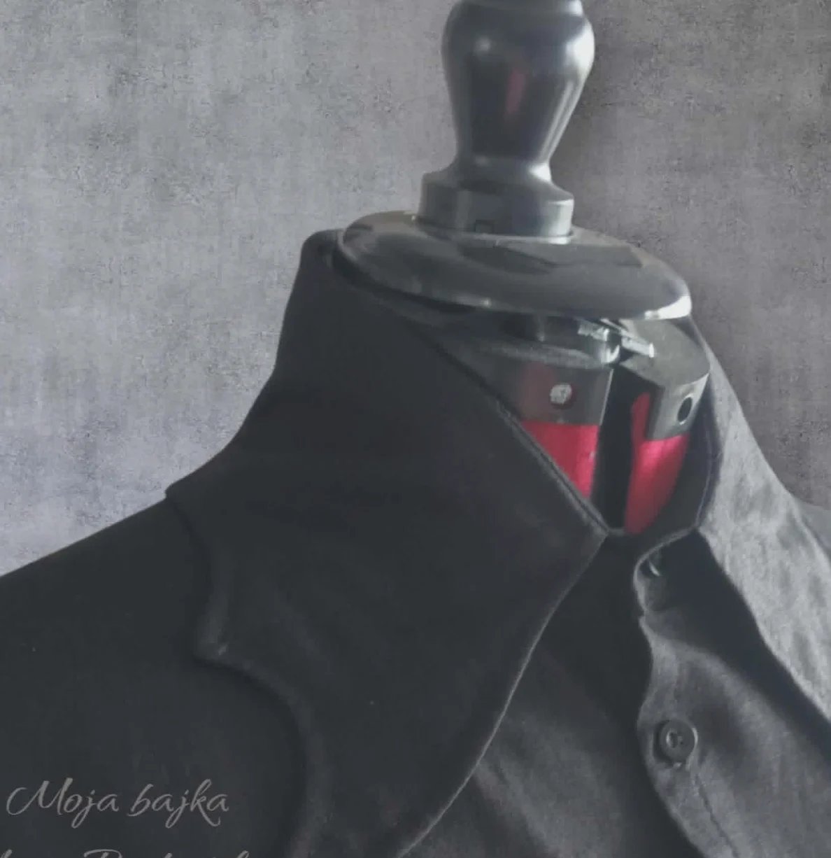 Mannequin wearing black coat draped with dark fabric and red geometric accessory against textured gray wall