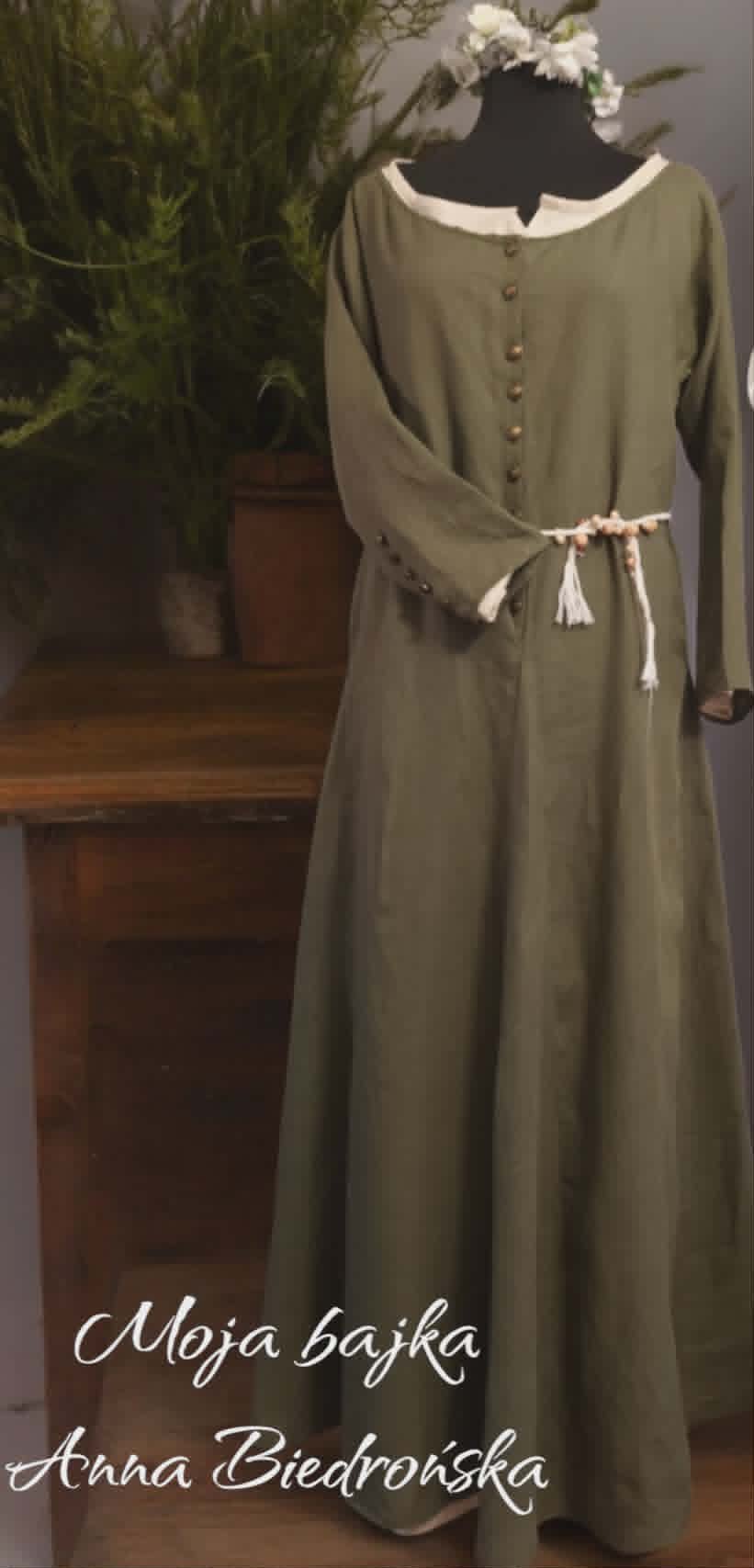 Mannequin wearing a sage green medieval-style dress with white collar and decorative cord belt, displayed next to a potted plant