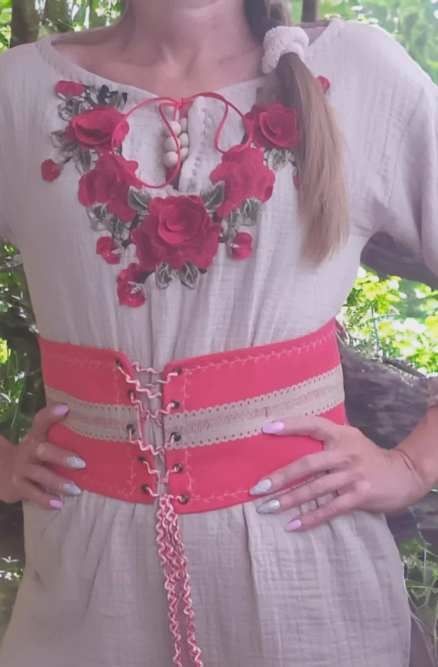 Woman wearing white peasant blouse with red rose embroidery and decorative pink belt with chain detail outdoors