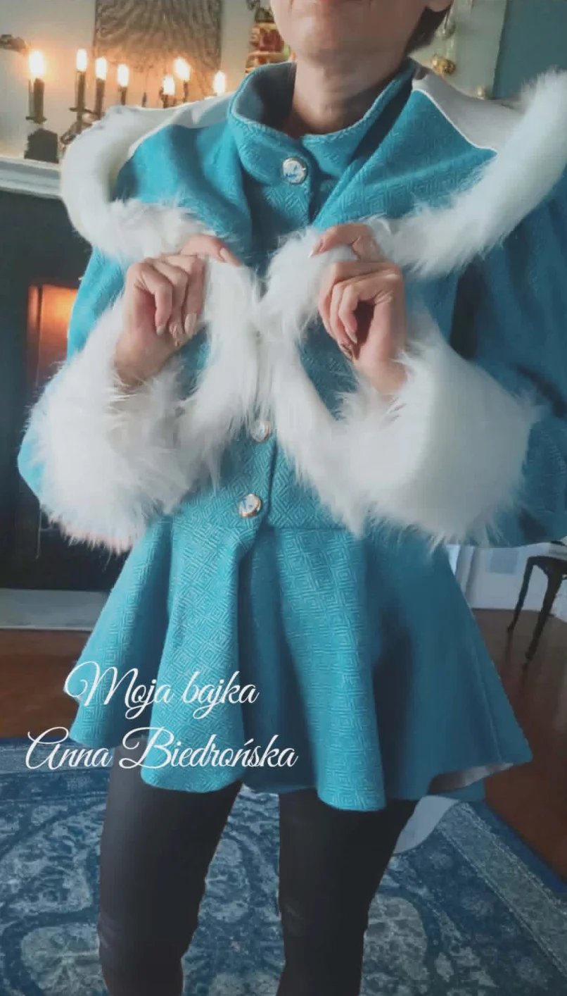 Person wearing a turquoise coat with white fur collar and cuffs, posing indoors with candles visible in the background
