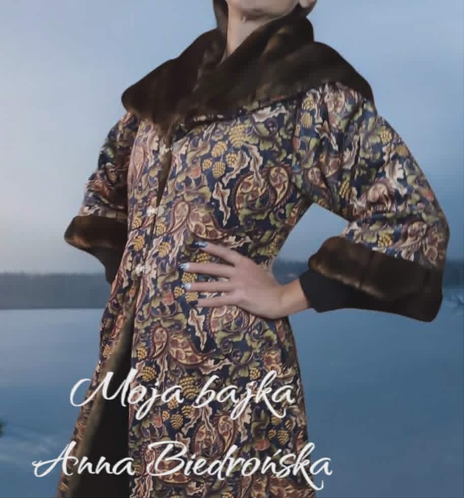 Woman wearing ornate patterned coat with dark fur collar standing by water, overlaid with white text credits