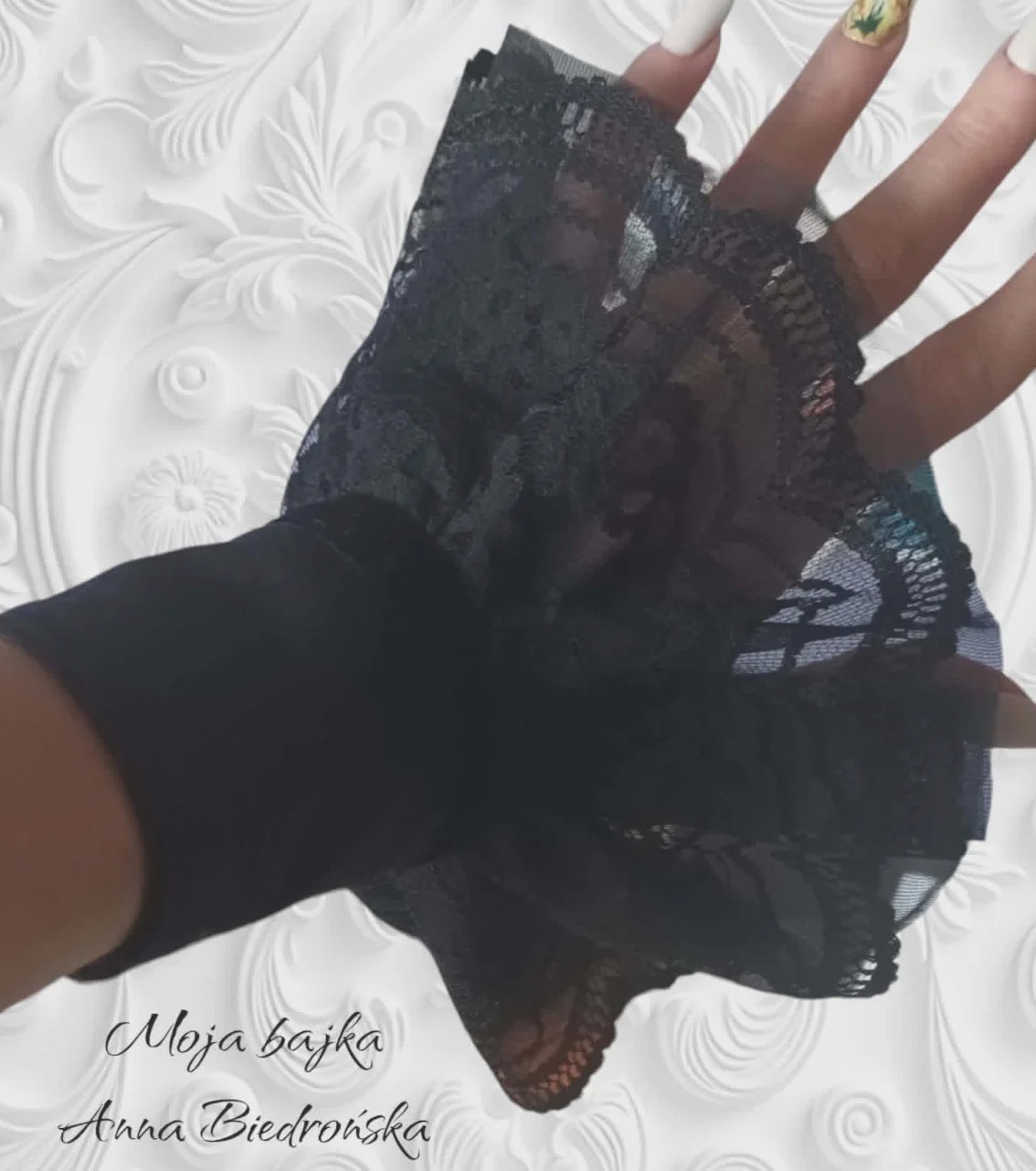Person wearing black lace fingerless gloves with embroidered details, displaying them on a white textured background