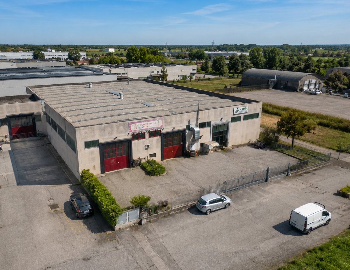 Aerial view of a large industrial warehouse with red garage doors, surrounded by a paved lot with parked vehicles and nearby green fields
