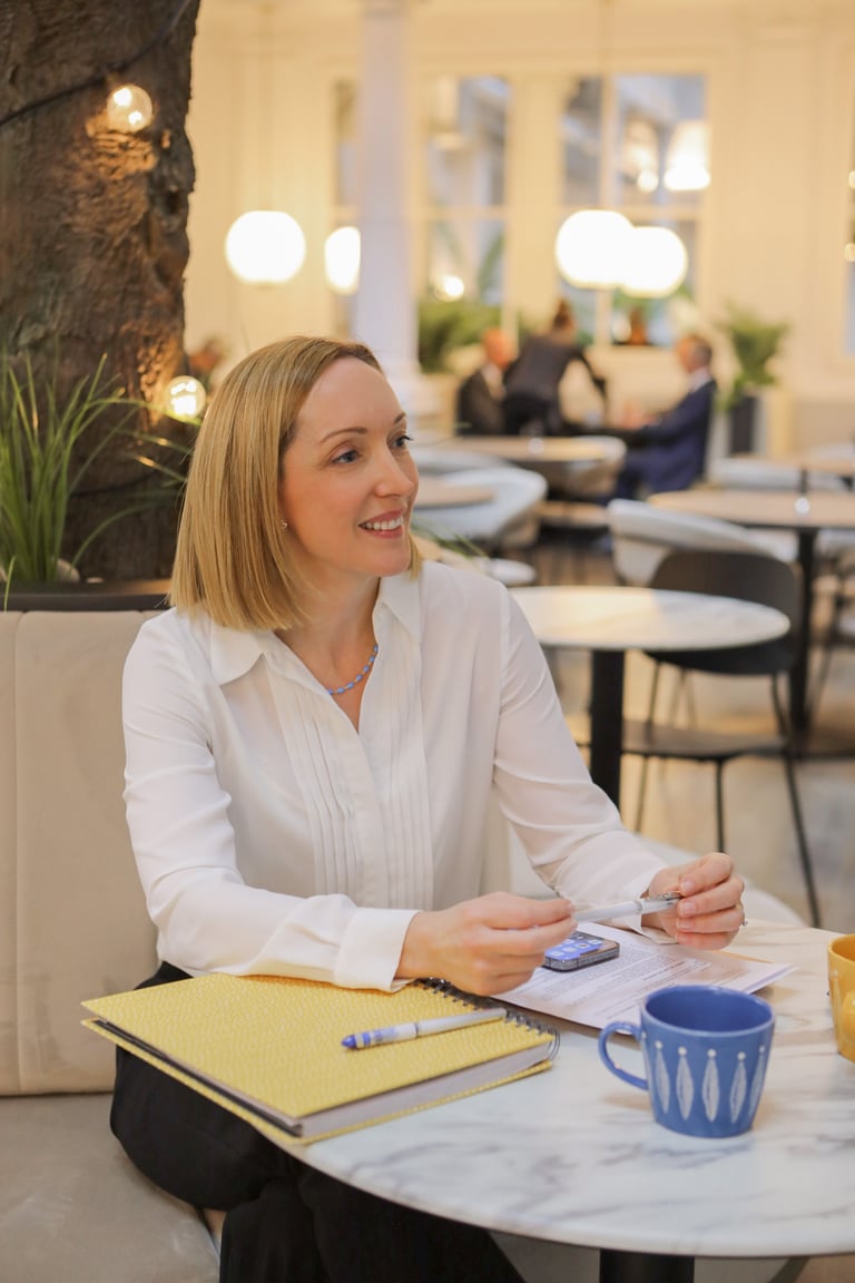 Woman in white blouse smiling at table with notebook and coffee mug in modern office setting