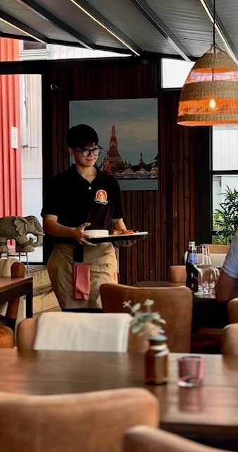 Restaurant server holding a plate in a modern dining room with dark wood interior and Bangkok temple view through window
