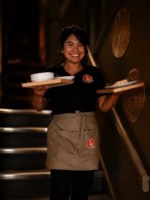 Smiling server in black shirt and tan apron holding wooden trays with food and dishes in a restaurant with decorative plates on the wall