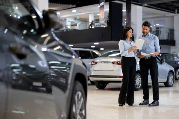 A man and woman in business casual attire discuss a vehicle in a modern car showroom with multiple cars visible in the background.