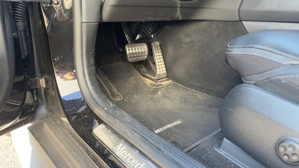 Interior view of a cars driver-side floor showing carpet, pedals, and door sill in daylight