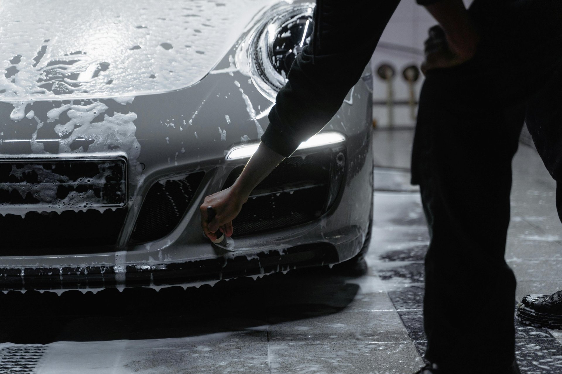 Person hand-washing the front bumper and headlight of a black car with soap and water outdoors
