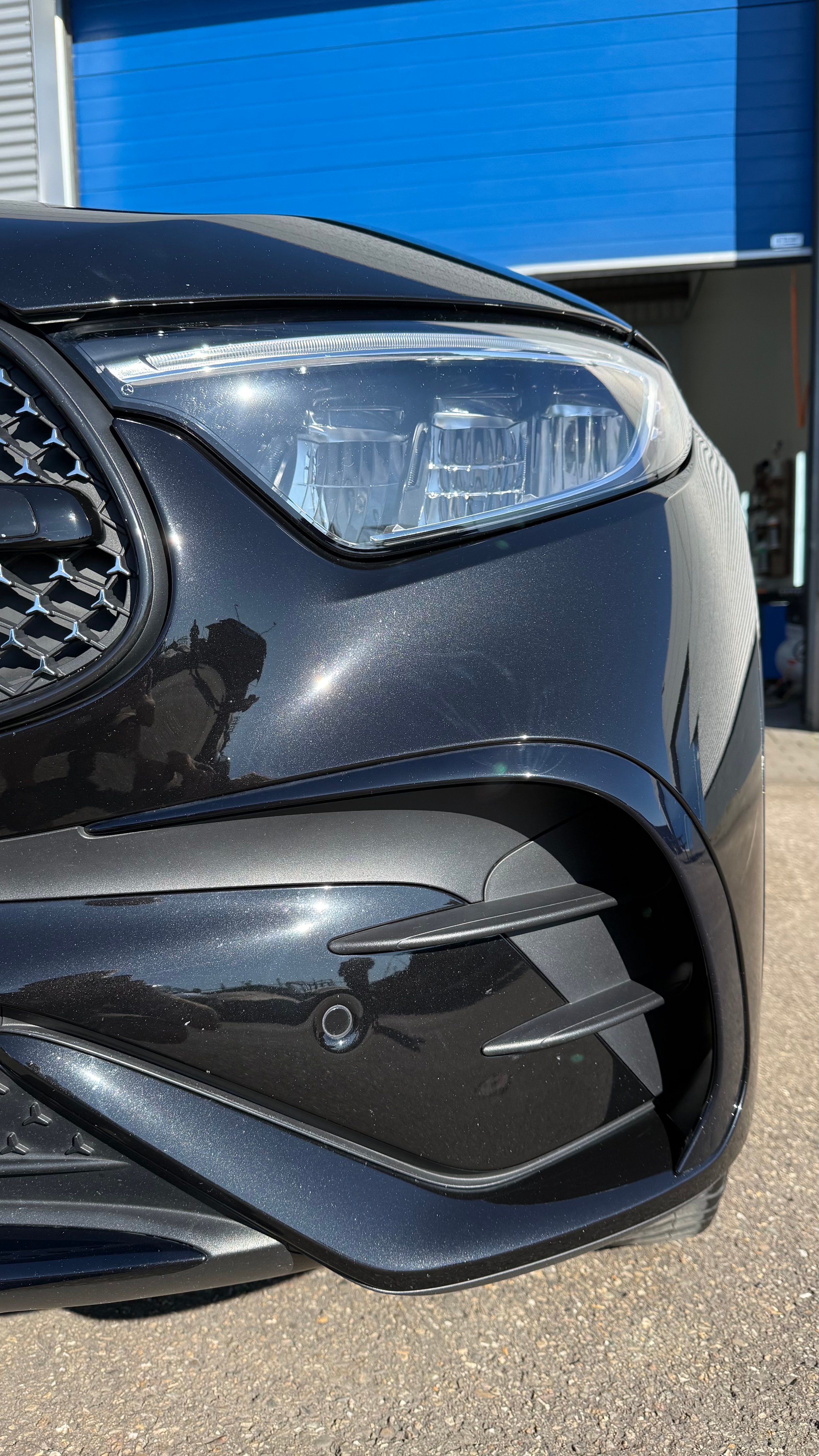 Na: Close-up of dark gray car front with LED headlight and lower air intake vent in outdoor parking area
