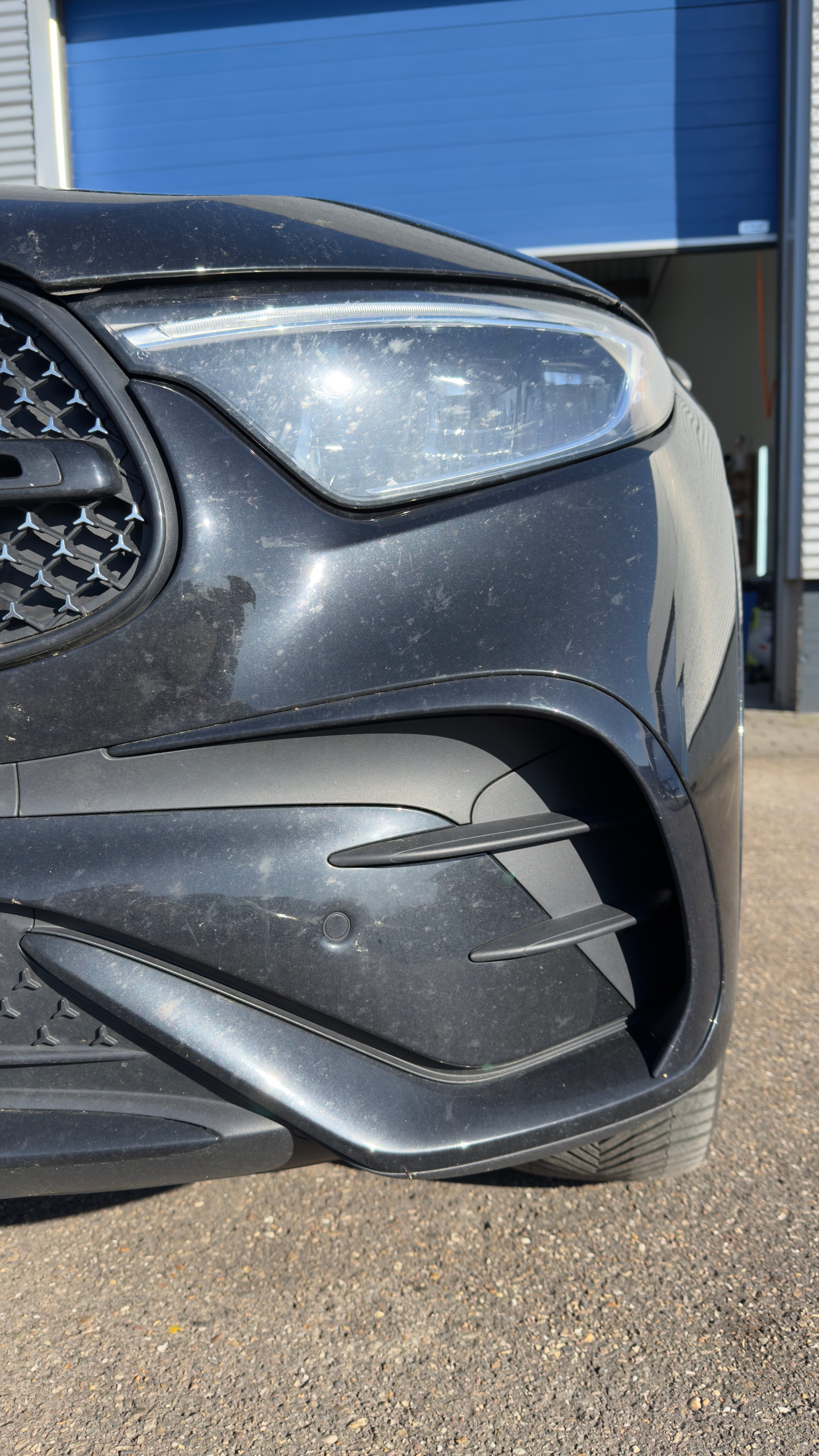 Voor: Close-up of a dark-colored cars front headlight and fender with dust buildup, parked near a blue building