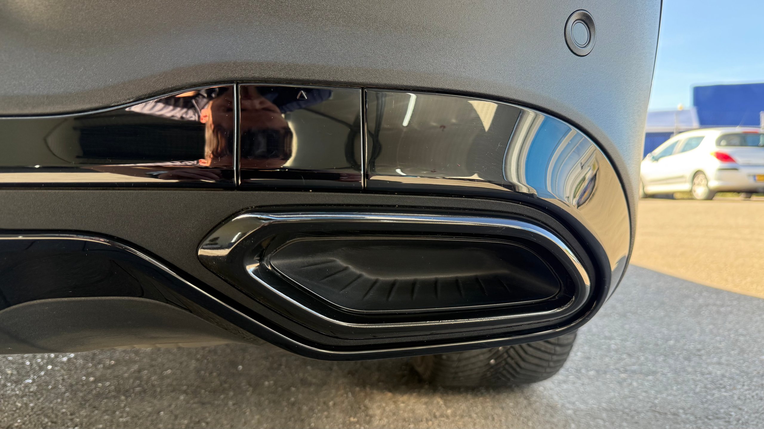 Na: Close-up of a sleek black car exhaust pipe with oval opening and chrome detailing on a gray road