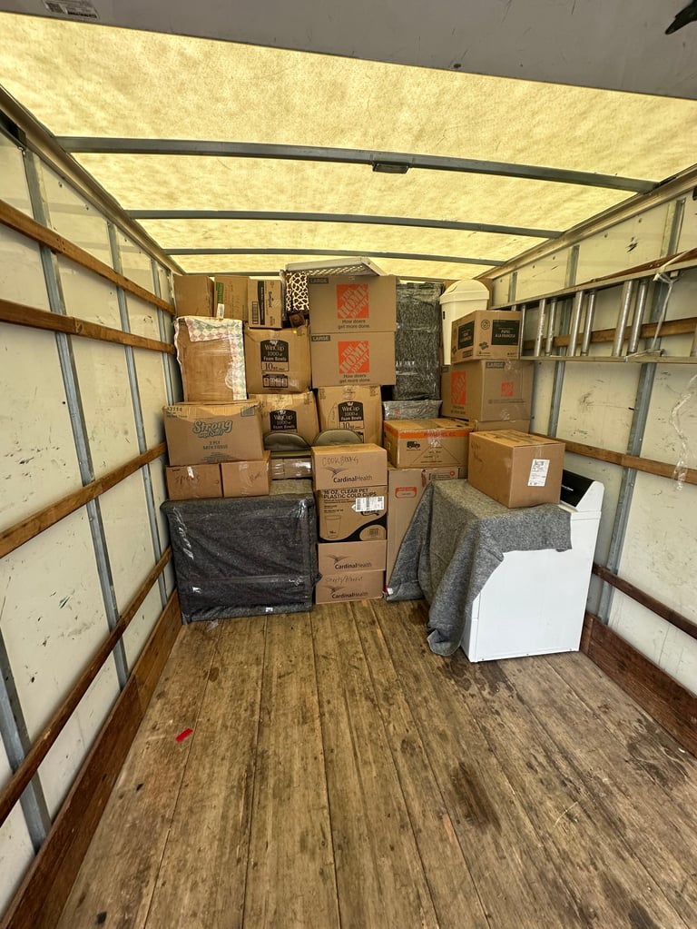 Interior of a moving truck or storage unit filled with stacked cardboard boxes, rolled textiles, and packed items on a wooden floor