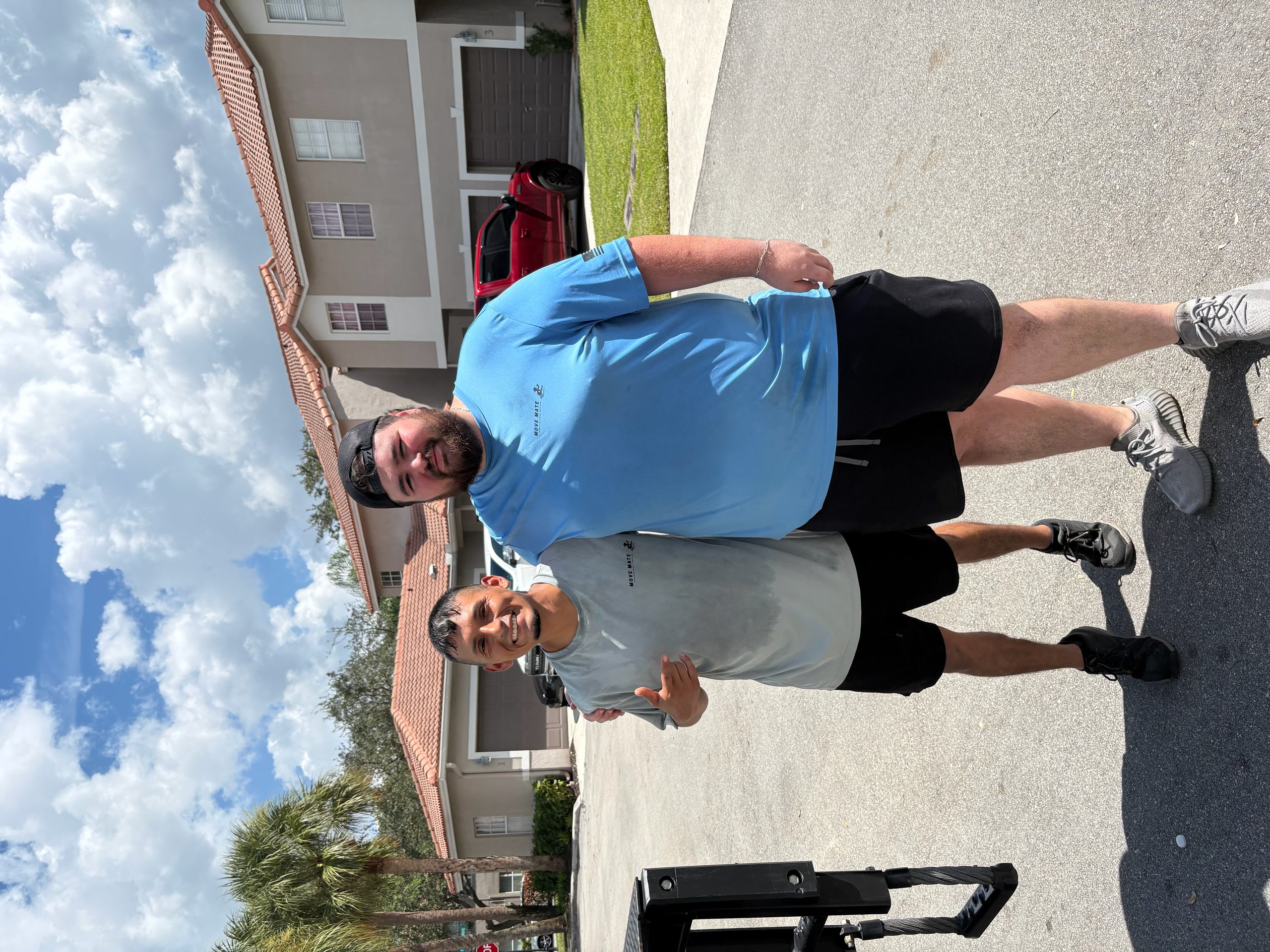Two men standing outdoors on a sidewalk, one wearing a blue shirt and the other wearing a black shirt, both smiling at the camera