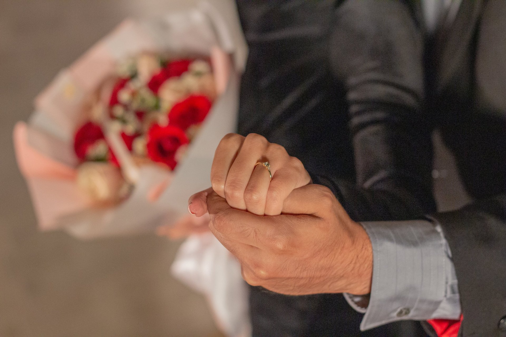 Couple holding hands with engagement ring, woman holding red rose bouquet in background