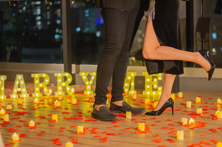 Two people standing among glowing candles and rose petals with illuminated MARRY ME letters for a marriage proposal