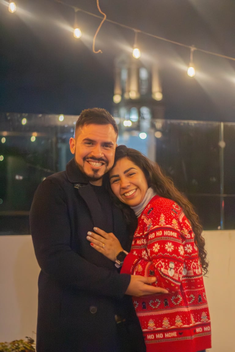Couple smiling at camera on rooftop at night with string lights and city skyline in background
