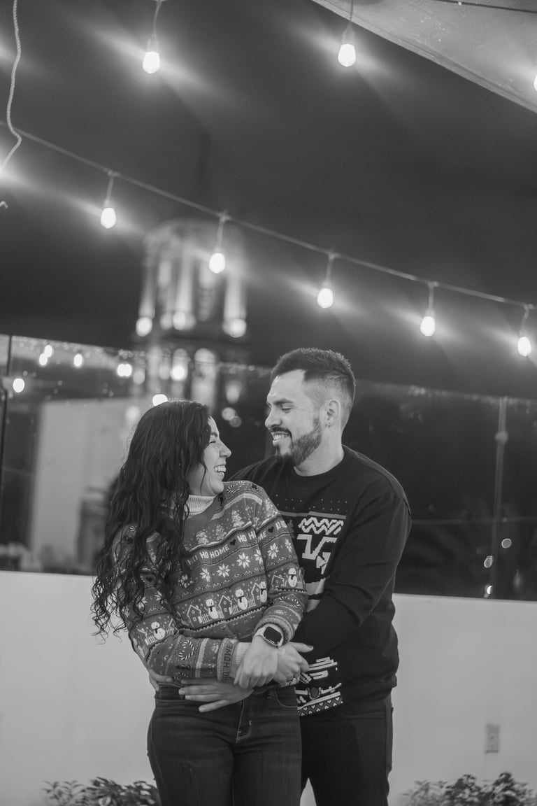 Couple in festive sweaters smiling together under string lights at night in black and white