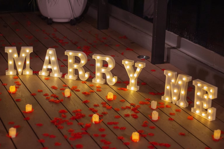 Illuminated MARRY ME letters on wooden floor with red rose petals and glowing candles for romantic proposal setup