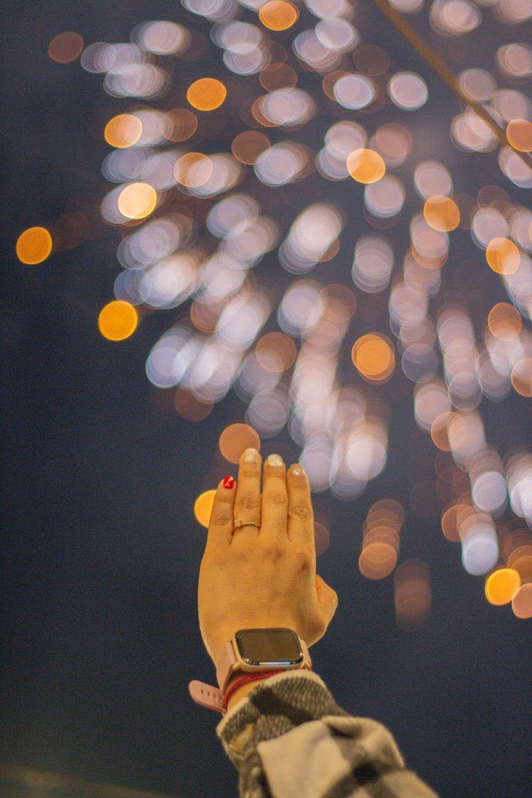 Hand raised toward bokeh lights at night, wearing red nail polish and a watch with warm glowing orbs in background