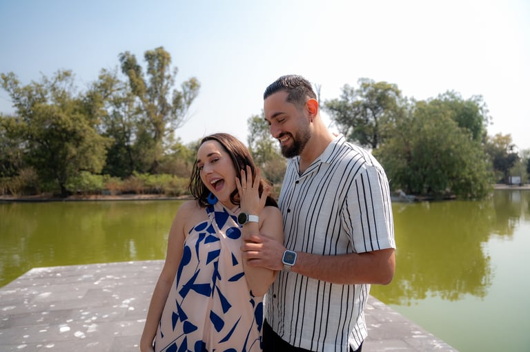 Couple smiling by a river with green trees and clear sky in the background. Woman shows engagement ring to camera.