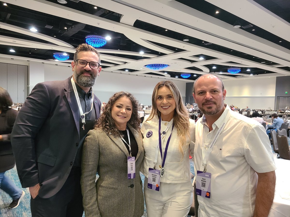 Four professionals wearing ID badges pose together at a conference or event in a modern venue with blue-lit ceiling panels