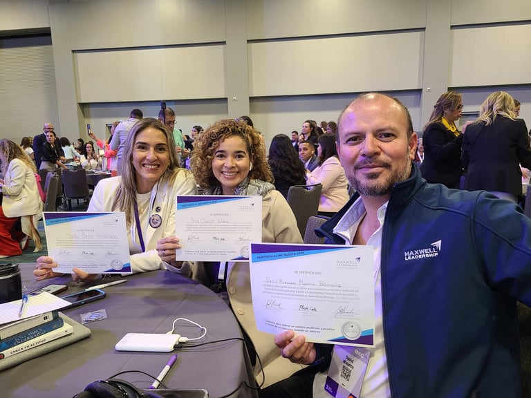 Three people holding certificates at an indoor event with attendees in background
