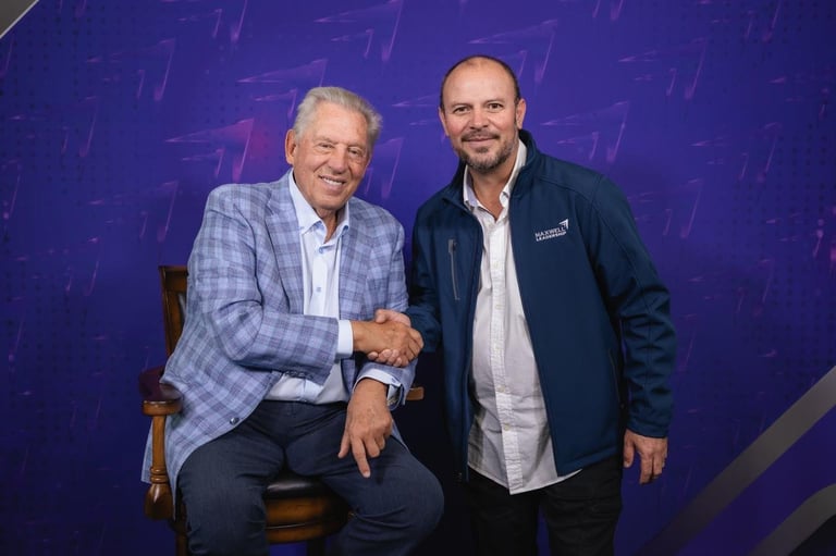 Two men in business casual attire shake hands while posing together against a purple textured background