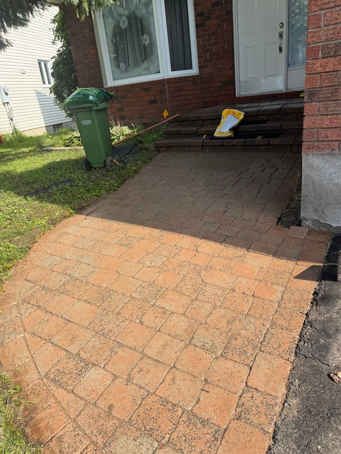 Residential driveway with red brick patio in foreground, dark asphalt in back, brick house with green trash bin, and white door visible