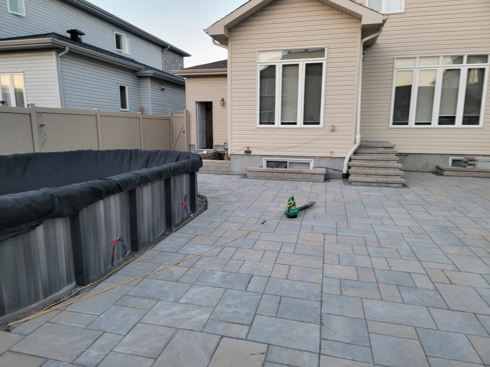 Residential backyard patio with gray pavers, beige house, metal fence, and steps leading to back entrance