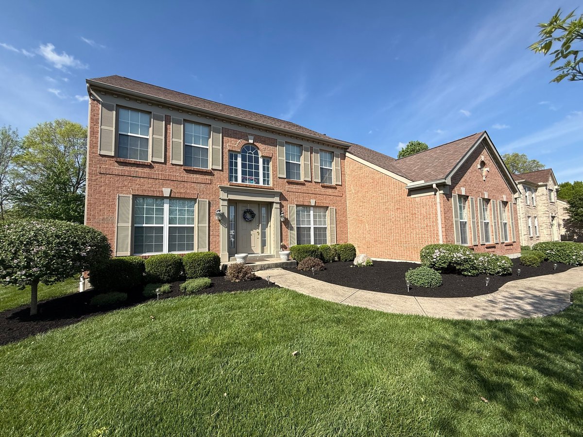 Two-story red brick residential home with manicured lawn, curved driveway, and landscaped shrubs under blue sky
