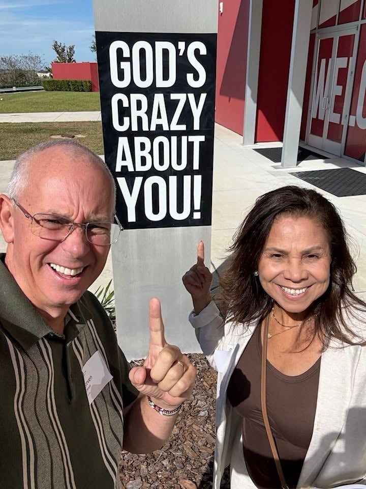 Two people smiling and making peace signs in front of a church sign reading Gods Crazy About You