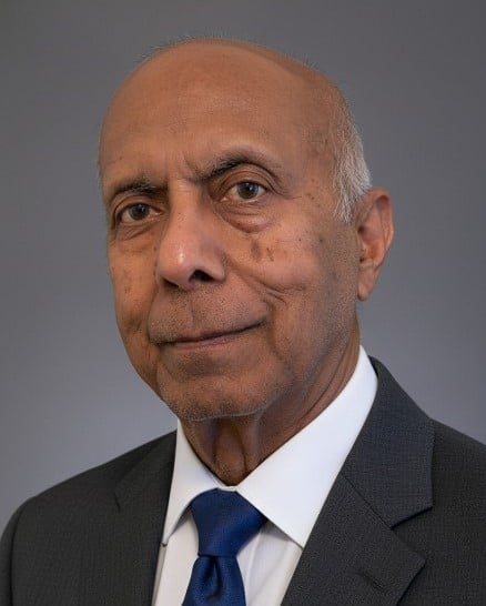 Professional headshot of a middle-aged man wearing a dark suit, white dress shirt, and navy blue tie against a gray background.