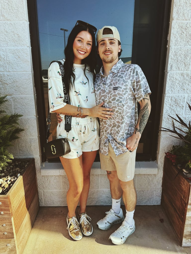 A smiling couple posing together at a doorway, the woman in a light floral top and the man wearing a patterned shirt and white cap