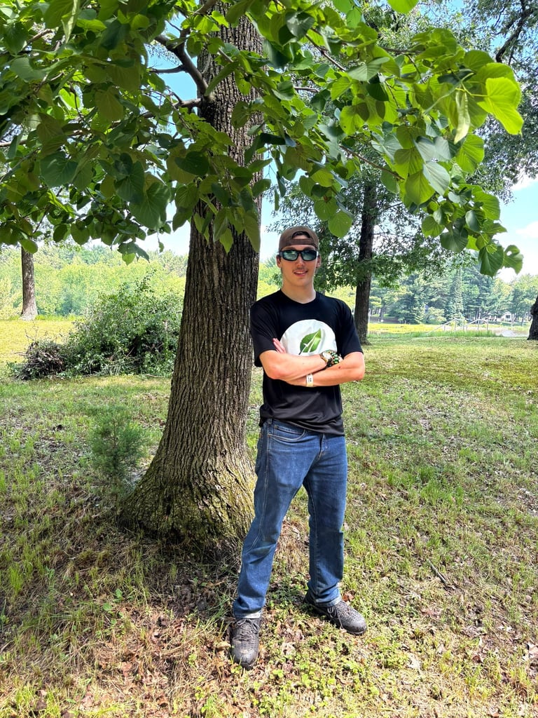 Man in black shirt and jeans standing under large tree in park, arms crossed, wearing sunglasses and hat
