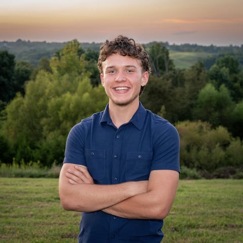 Smiling young man in navy shirt with arms crossed standing in front of rolling countryside and forest landscape at sunset