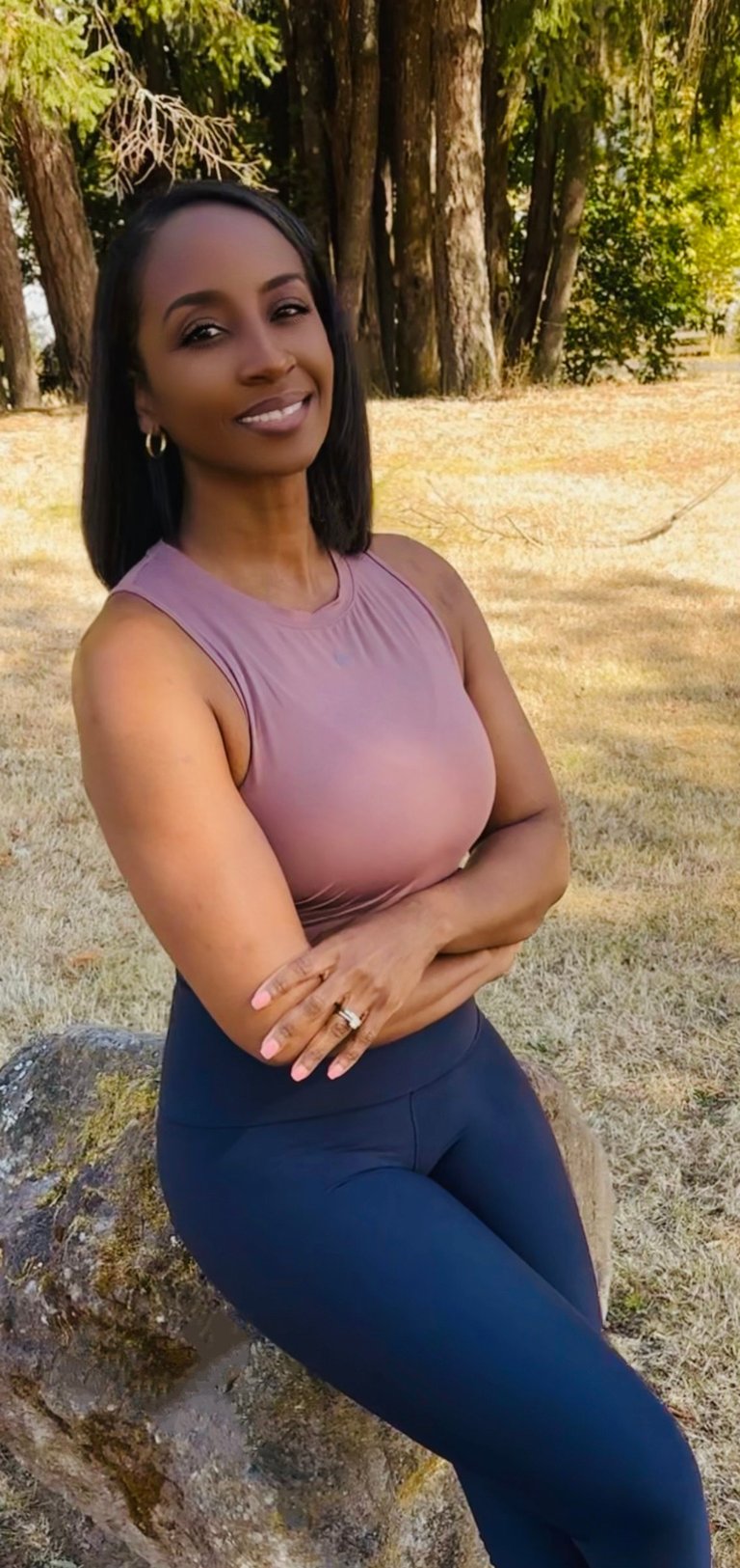 Natari smiling at camera wearing a pink sleeveless top and navy leggings, posing outdoors near trees and dry grass