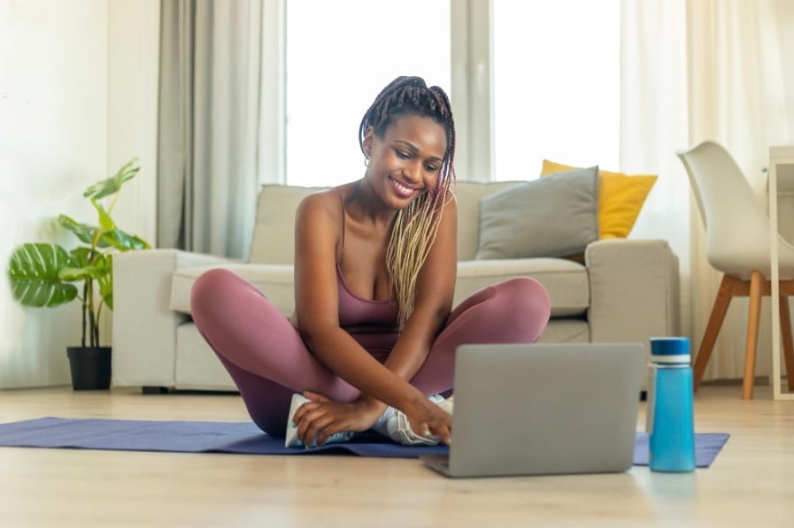 Natari in workout clothes sitting on yoga mat using laptop in bright living room with couch and plants