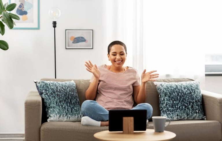 Woman sitting on a beige couch gesturing enthusiastically while looking at a tablet in a bright, modern living room