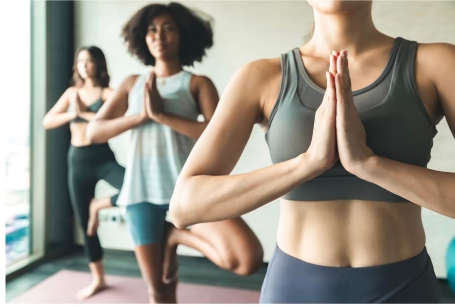 Group of women in fitness attire practicing yoga indoors, with one woman in front doing a prayer pose position