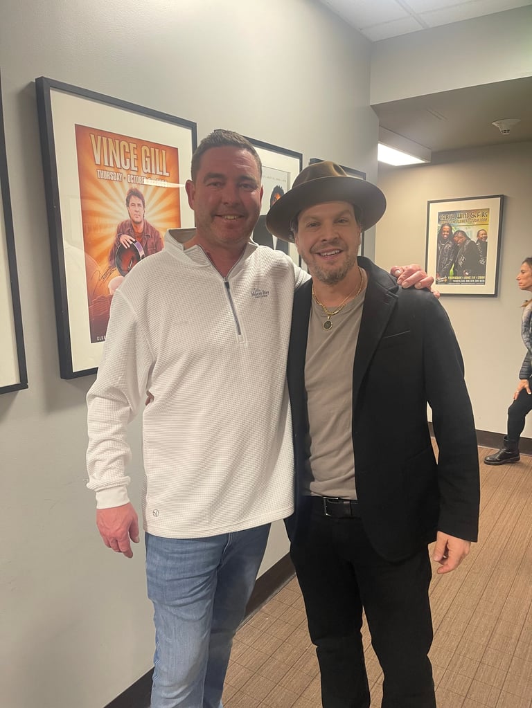 Two men posing together indoors, one wearing a white jacket and jeans, the other in a black blazer and hat, with movie posters visible in the background
