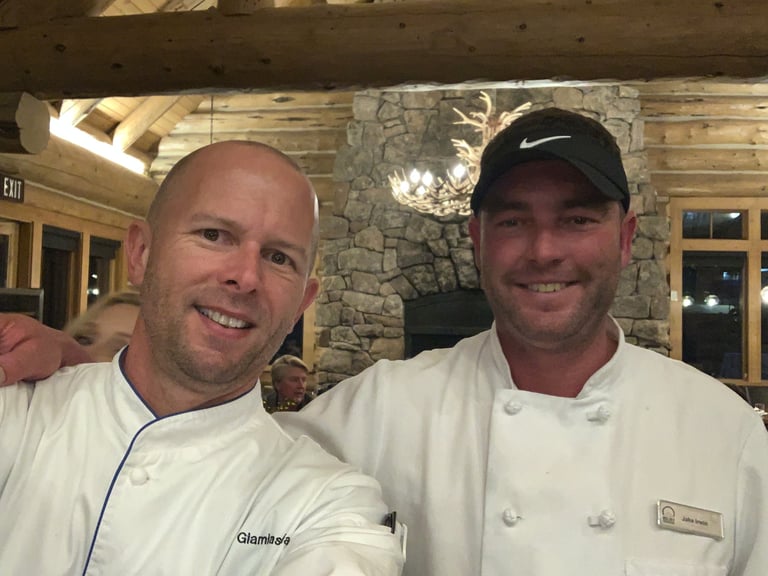 Two men in white chef coats smiling at camera in front of a stone fireplace and wooden beams