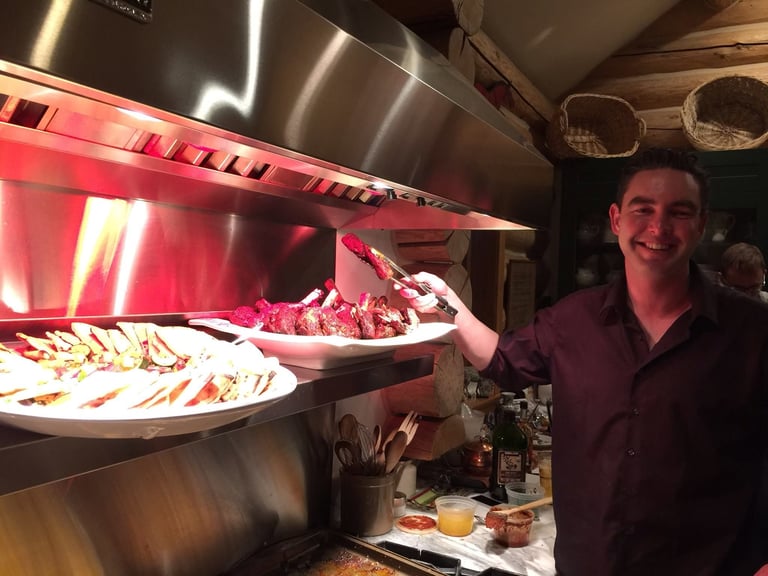 Man in purple shirt smiling while working in commercial kitchen with red heat lamps and plated dishes