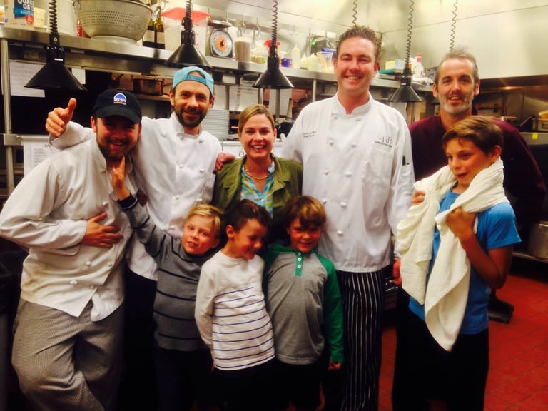 Group of adults and children posing together in a professional kitchen, smiling at the camera