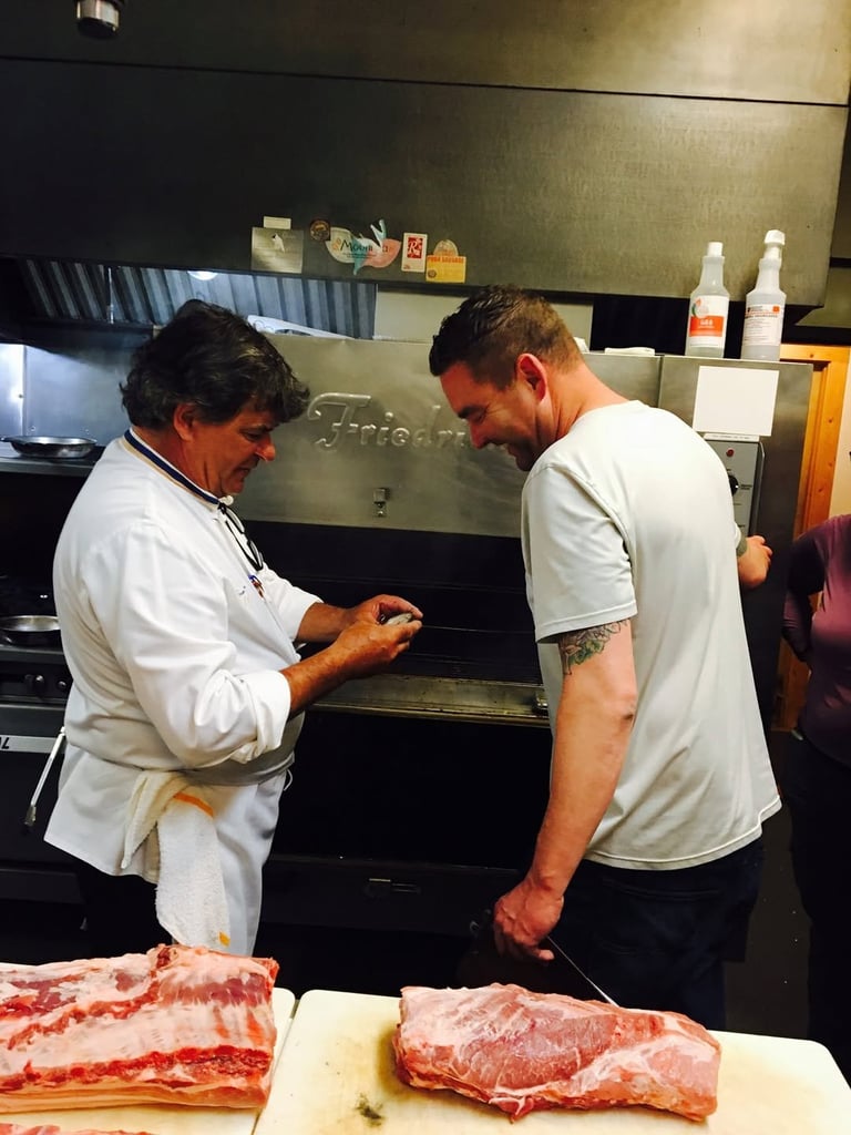 Two men examine meat at a kitchen counter near a commercial oven in what appears to be a butcher shop or restaurant kitchen